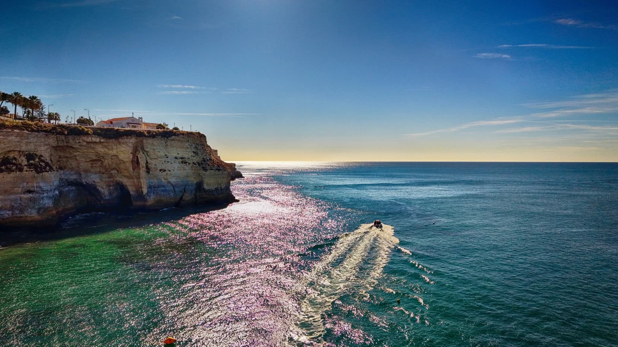 A small boat cutting through a glittering, sunlit sea below Carvoeiro's cliffs
